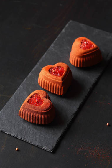 當(dāng)代巧克力迷你慕斯蛋糕由心臟形狀硅膠模具制成，覆蓋著磚色天鵝絨噴霧，并裝飾有血橙魚子醬，黑色背景