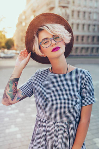 竖线肖像漂亮的金发女孩与短发摆姿势的相机在城市的骏马。她穿着灰色格子裙，眼镜，帽子，有紫色的嘴唇，纹身。她看着镜头