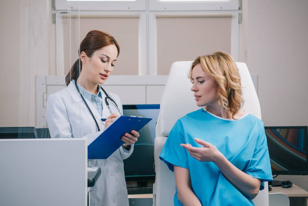 漂亮的医生写在剪贴板,而站在病人坐在扶手椅附近 漂亮的医生写在剪贴板,而站在病人坐在扶手椅附近