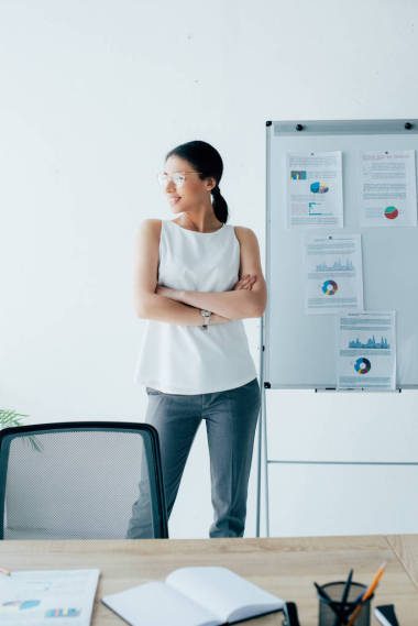 有吸引力的拉丁女商人站在交叉的手臂附近的活動(dòng)掛圖，并在辦公室望去