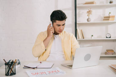 細(xì)心的亞洲男子在智能手機(jī)上交談,一邊坐在家里的辦公桌前,一邊使用筆記本電腦