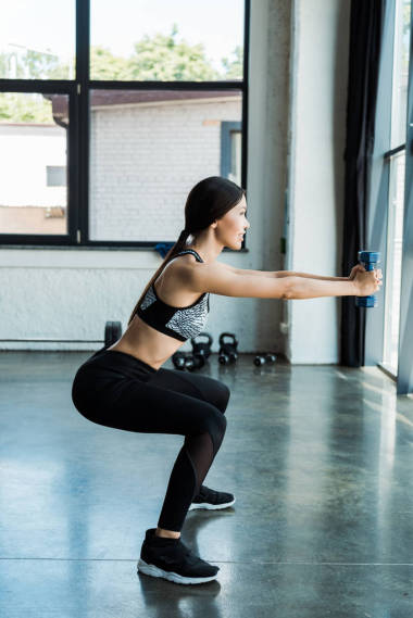 側(cè)看快樂的年輕女運(yùn)動(dòng)員拿著啞鈴和做蹲下運(yùn)動(dòng) 