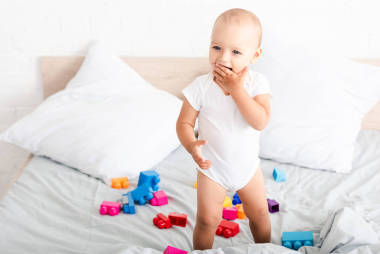 微笑赤腳嬰兒在白色的衣服站在床上與玩具，并采取手指到他的嘴