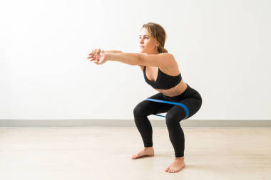 相當(dāng)自信的年輕女性黑發(fā)做蹲與阻力帶對(duì)白墻的圈