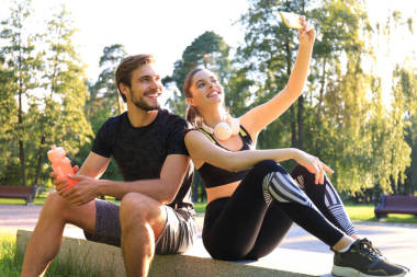 身穿运动服的年轻夫妇坐在城市公园的栏杆上，在夏日阳光灿烂的日子里锻炼完后自拍