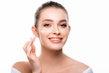 迷人的、面帶微笑的女人在臉上涂化妝品，看著被白色隔離的相機(jī)