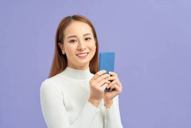 微笑可愛的年輕女子站在紫羅蘭色背景下使用手機(jī)