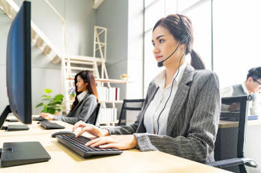 年輕、友善、自信的接線員女探員與同事在呼叫中心工作的耳機(jī)一起微笑，背景是客戶服務(wù)和技術(shù)支持工作場(chǎng)