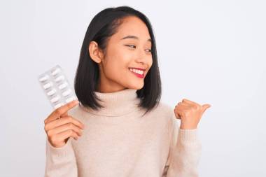 年輕美麗的中國女人拿著藥丸，站在孤立的白色背景上，用拇指指向旁邊，笑容滿面