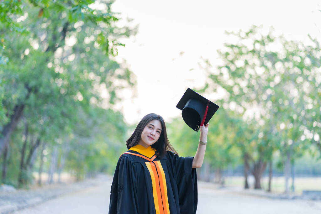 一位穿着学术礼服头戴研究生帽的年轻快乐的女毕业生在大学校园里开始庆祝她的大学学位