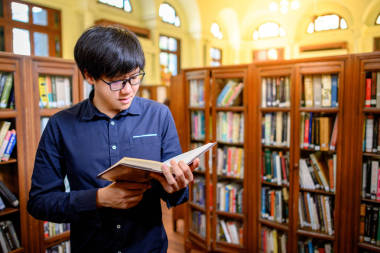 聰明的亞洲人大學(xué)生戴著眼鏡，坐在老式書(shū)架上看書(shū)。高校圖書(shū)館的教材資源用于教育科目和研究.教育機(jī)會(huì)獎(jiǎng)學(xué)金