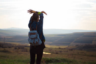 在日落和田野的背景下，穿著背包斜紋棉布夾克，手舉著手的年輕女孩的背景肖像