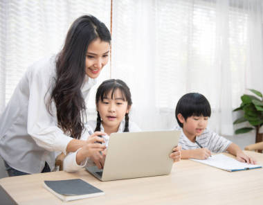 在家里客廳里，一位年輕的亞洲女人坐在桌子旁邊，用筆記本電腦幫她兒子和女兒做作