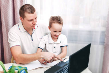 家庭远程在线学习。爸爸和儿子坐在窗边的桌子旁，在笔记本电脑上看视频课。文字的位置。检疫验尸官。父亲