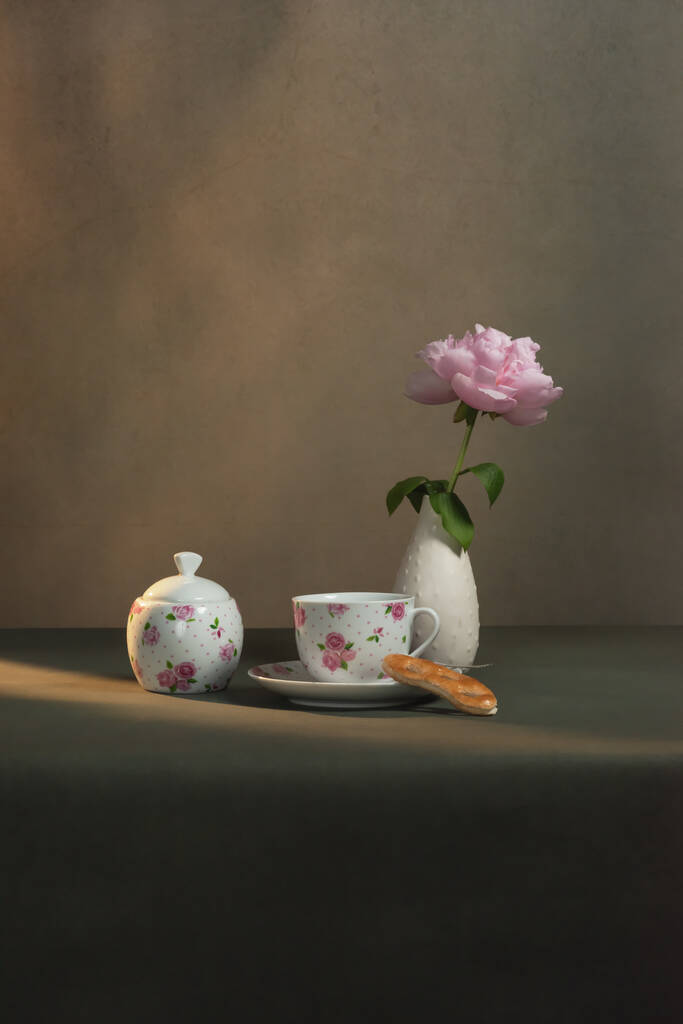 茶杯，茶碟和曲奇饼，糖碗，白色花瓶，还有牡丹，放在清晨阳光下灰色房间的桌子上