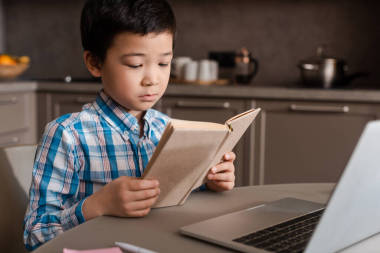在自我隔離期間，亞洲男孩在家里看書(shū)，帶著筆記本電腦上網(wǎng)學(xué)習(xí)