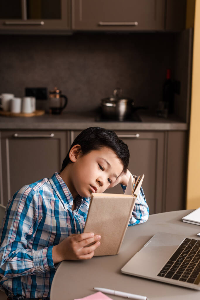 在自我隔离期间,亚洲小孩在家里看书,带着笔记本电脑上网学习 在自我隔离期间,亚洲小孩在家里看书,带着笔记本电脑上网学习