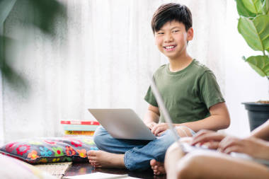 聰明的亞洲少年使用電腦筆記本電腦，坐在木制地板上，笑著，與朋友討論小組工作、家庭作業(yè)、考試準備等問題。小組工作與協(xié)作