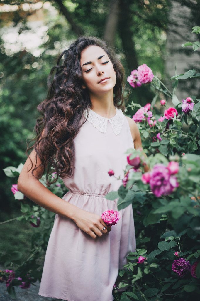 美丽的年轻女子,穿着粉红色的连衣裙摆在一个玫瑰花园 美丽的年轻女子,穿着粉红色的连衣裙摆在一个玫瑰花园