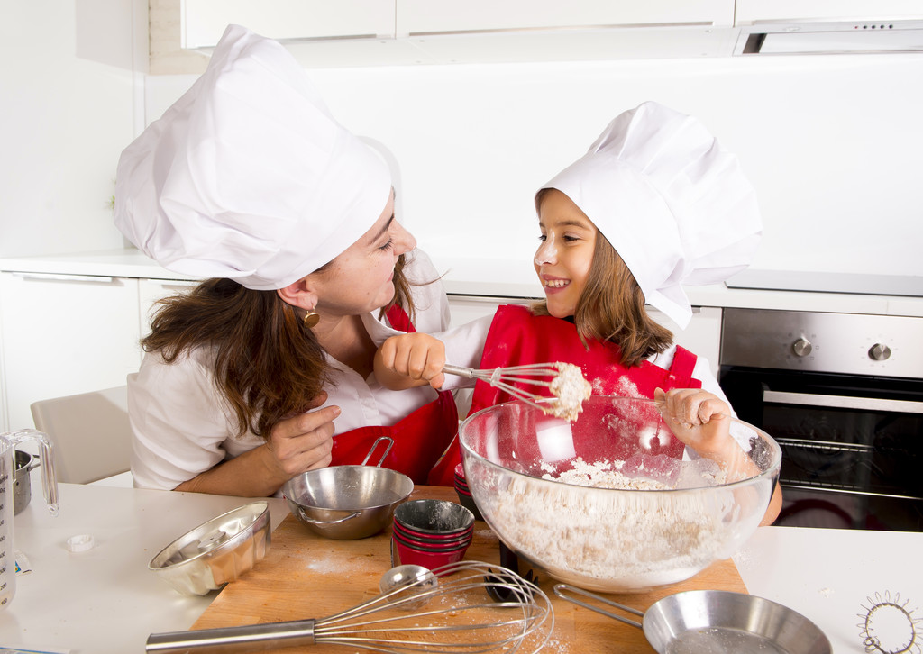 快乐的妈妈和穿着围裙的小女儿在厨房用面粉做帽子 快乐的妈妈和穿着围裙的小女儿在厨房用面粉做帽子